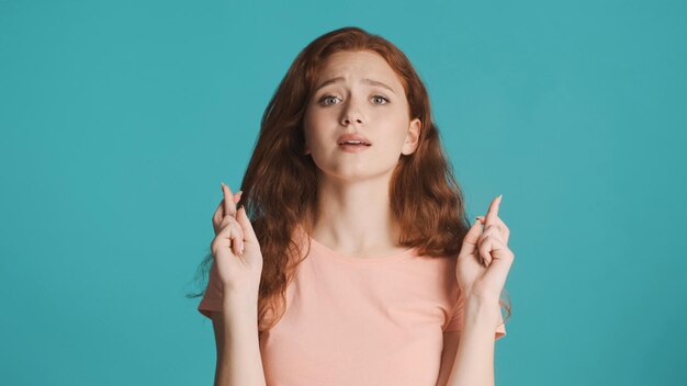 Emotional redhead girl with crossed fingers hopefully praying on camera over colorful background Please expression