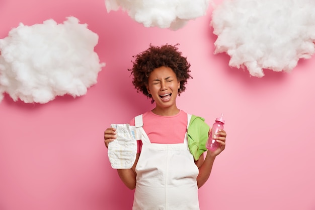 Free photo emotional sad pregnant woman cries with despair, worries as doesnt have experience of motherhood, holds nappy and feeding bottle, tired of pregnancy, being on third trimester, isolated on pink wall