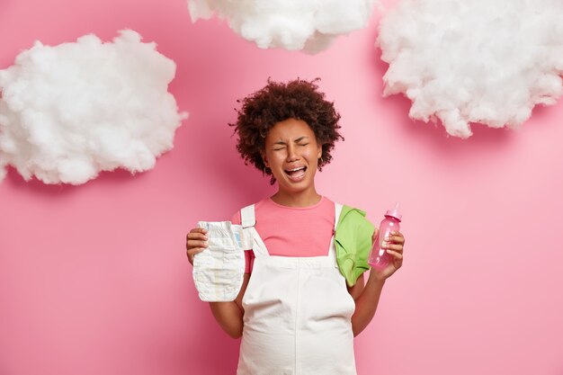 Emotional sad pregnant woman cries with despair, worries as doesnt have experience of motherhood, holds nappy and feeding bottle, tired of pregnancy, being on third trimester, isolated on pink wall