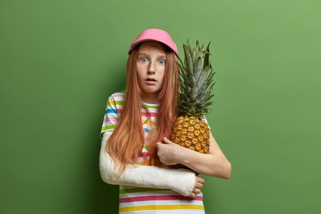 감정적으로 무서워하는 주근깨가있는 소녀는 파인애플을 포용하고 열대 과일을 좋아하며 모자와 줄무늬 티셔츠를 입고 녹색 벽에 고립 된 팔이 부러졌습니다. 어린 시절과 라이프 스타일 개념
