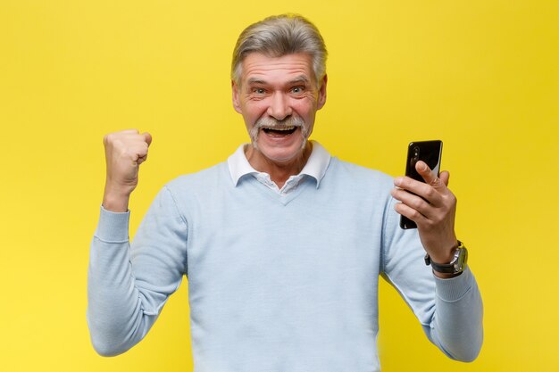 Эмоциональный старший мужчина с телефоном выигрывает что-то, позируя на желтой стене