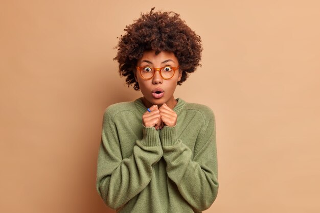 Emotional shocked woman with curly hair holds breath keeps hands together opens mouth with amazement stares bugged eyes dressed in casual jumper and optical spectacles models indoor