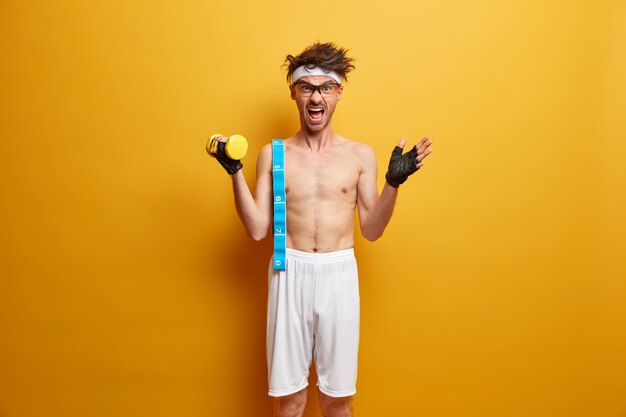 Emotional sportsman lifts heavy dumbbell, shouts emotionally, carries measuring tape, wears sport gloves, leads healthy lifestyle, poses against yellow wall. Body care, sport and diet concept
