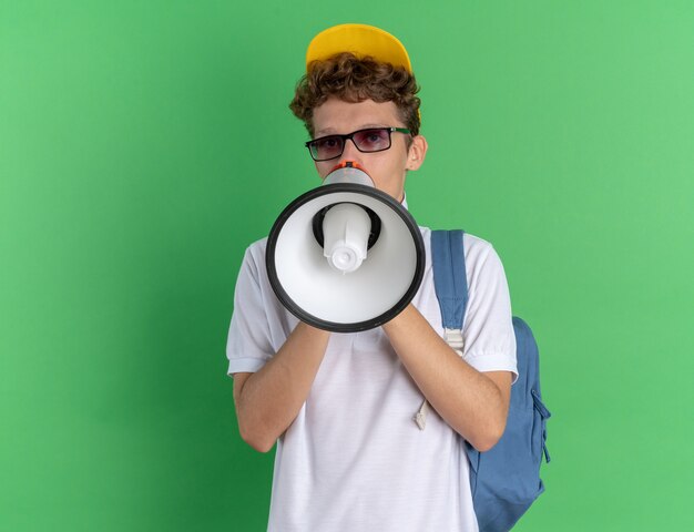 Emotional student guy in white polo shirt and yellow cap wearing glasses with backpack shouting to megaphone standing over green background
