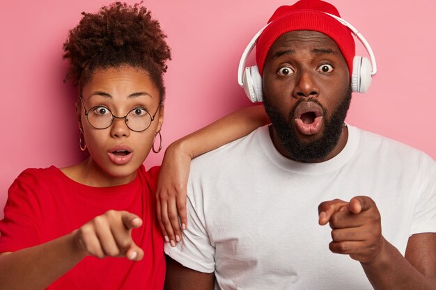 emotional stupefied ethnic young boyfriend and girlfriend point at camera, notice something unbelievable, have scared gazes, man wears stereo headphones on ears. Omg concept