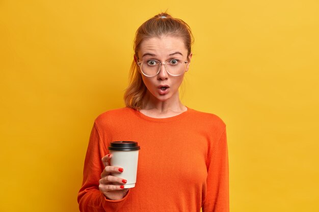 Emotional stupefied female student has coffee break, holds disposable cup of cappuccino, wears big transparent glasses, orange sweater, hears fresh gossip about groupmate, drinks caffeine beverage