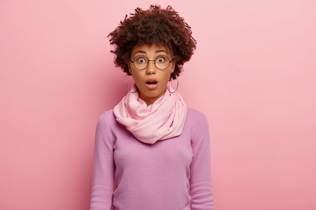 Emotional stupefied woman with curly Afro hairstyle, keeps mouth opened, wears round spectacles, casual jumper, shocked by puzzled news, opens mouth from wonder