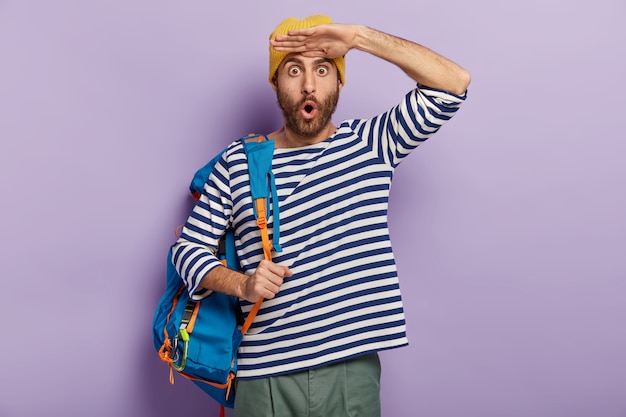 Emotional stupefied young man cannot believe in something, keeps palms near forehead, wears yellow hat and striped jumper