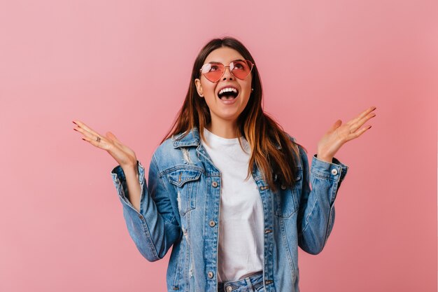 Emotional stylish woman looking up on pink background. Surprised brunette girl in denim jacket posing with open mouth.