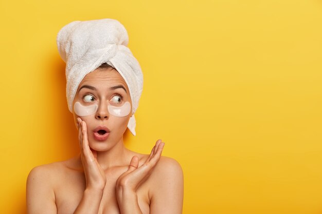 Emotional surprised Caucasian woman looks with shocked expression aside, stands bare shoulders against yellow wall, has flawless soft skin, wears patches under eyes, cleans her complexion