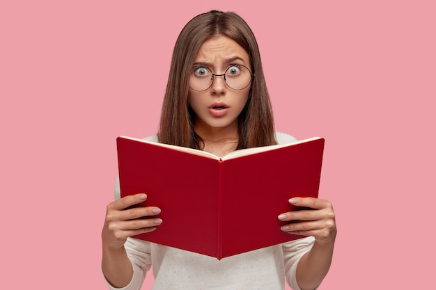 Emotional surprised European woman holds textbook, has scared facial expression, worries before passing entrance exam, wears round spectacles isolated over pink wall. Feminine girl with red book