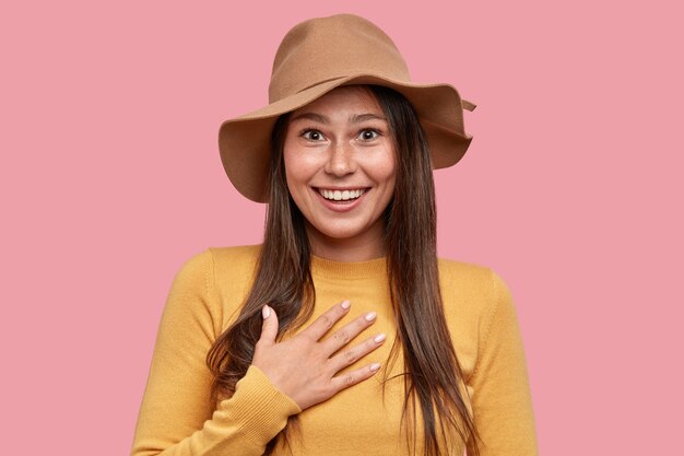 Emotional surprised freckled woman with positive expression keeps hand on chest, smiles broadly at camera