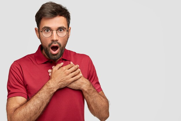 Emotional surprised male looks with bated breath, keeps mouth opened, terrific gaze can`t believe his eyes, wears casual red t-shirt, stands over white wall with blank copy space for text