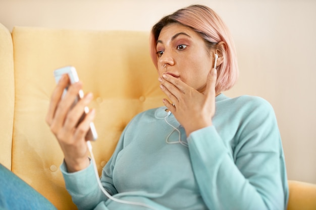 Emotional surprised young woman with pinkish hair sitting on sofa with smart phone, covering mouth in astonishment