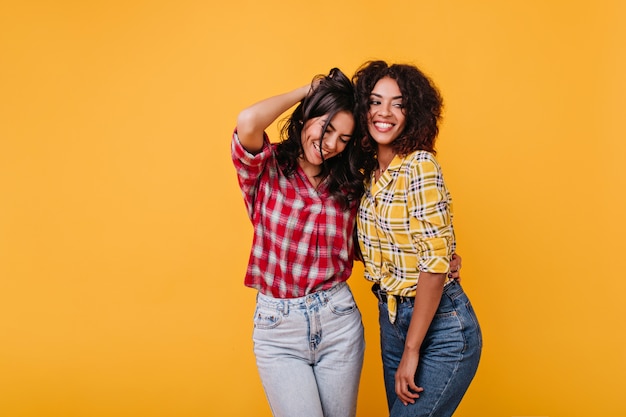 Emotional tanned women happily pose in plaid shirts. Portrait of brunettes with curly hair.