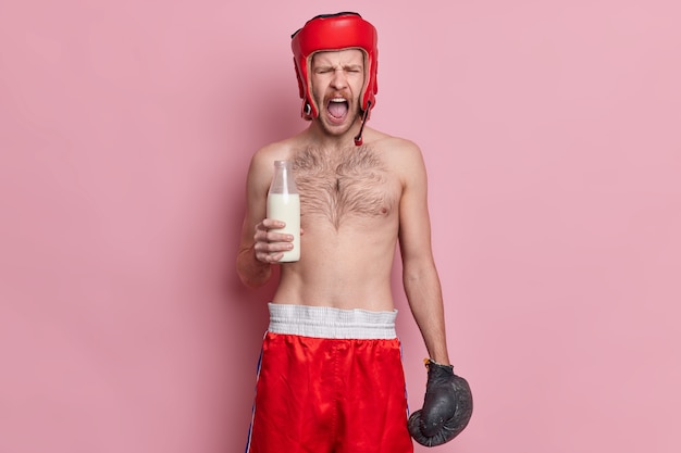 Free photo emotional topless male boxer shouts loudly keeps mouth opened holds glass bottle of milk wears hat shorts and boxing gloves.