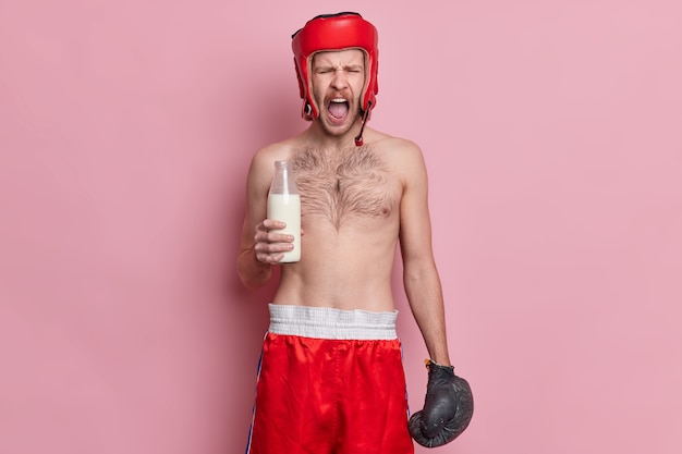 Emotional topless male boxer shouts loudly keeps mouth opened holds glass bottle of milk wears hat shorts and boxing gloves.