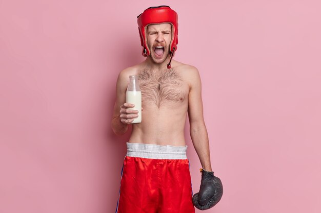 Эмоциональный мужчина-боксер топлес кричит, громко держит рот открытым, держит стеклянную бутылку с молоком, носит шляпу, шорты и боксерские перчатки.