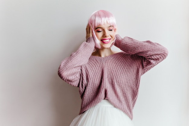 白い壁の前に立っている光沢のあるピンクの髪を持つ感情的なトレンディな女の子。 perukeと特大の紫色のセーターを着たのんきな女性の屋内写真。