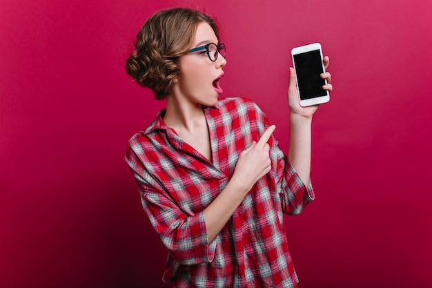 Emotional white girl with curly hairstyle posing with smartphone on claret wall. indoor portrait of amazed stylish woman in casual attire holding cell.