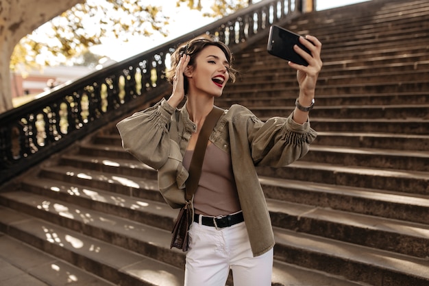 Emotional woman in denim jacket and white jeans making selfie. Curly woman with handbag taking photo outside.