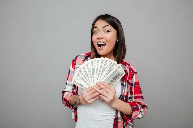 Emotional woman holding money over grey wall.