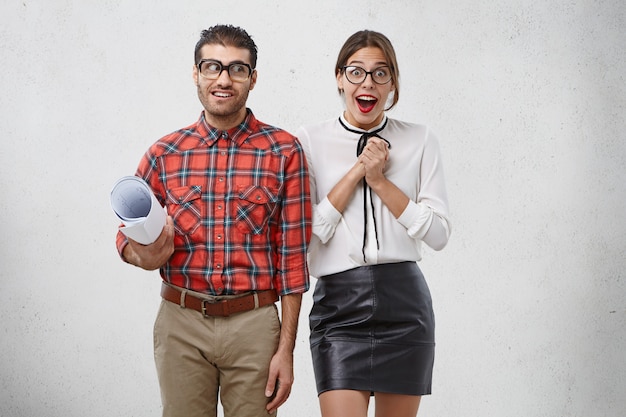 Emotional woman keeps hands together, stares at camera with astonishment stands next to male colleague who holds blueprint