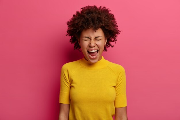 Emotional woman keeps mouth wide opened, yells and screams loudly, cannot control her emotions, being angry with someone, wears yellow t shirt, isolated on pink wall, reproaches someone