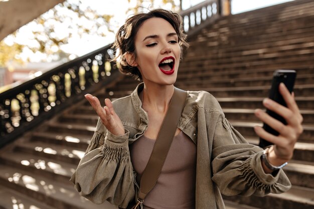 Emotional woman in long sleeve olive jacket making selfie outside. Cool woman with red lips taking photo