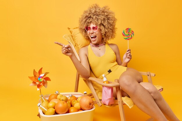 Emotional woman points away and shouts loudly dressed in swimsuit trendy pink sunglasses holds caramel candy on stick poses on deck chair against yellow background Summer time and rest concept