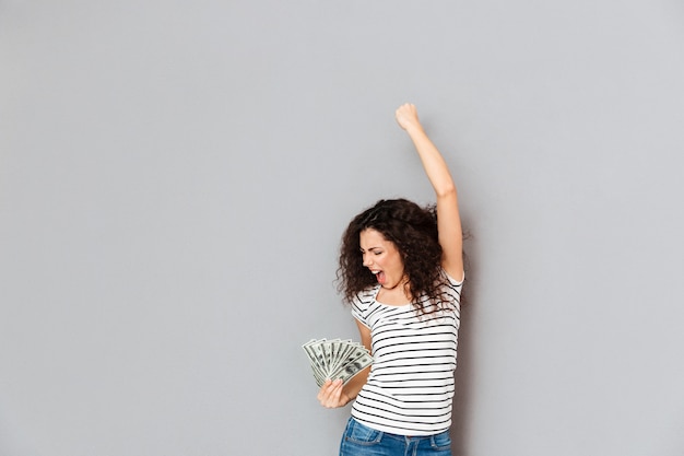 Emotional woman in striped t shirt acting like winner holding fan of 100 dollar bills and clenching fist in the air over grey wall