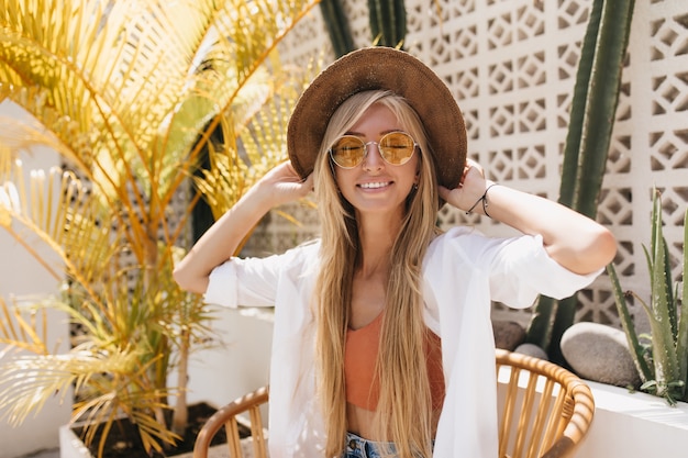 Emotional woman with blonde straight hair posing with eyes closed in resort restaurant. charming caucasian female model in brown hat smiling during photoshoot in cafe.
