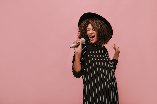 Бесплатное фото Эмоциональная женщина с кудрявой прической брюнетки в стильной шляпе и полосатом черном платье держит микрофон и поет на розовой стене.