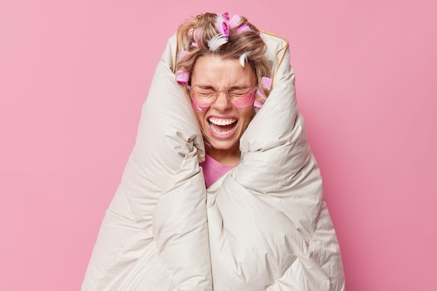 Free photo emotional woman yells loudly wrapped in soft blanket wears transparent eyeglasses hydrogel patches makes hairstyle with rollers poses in bedroom against pink background. morning daily routines