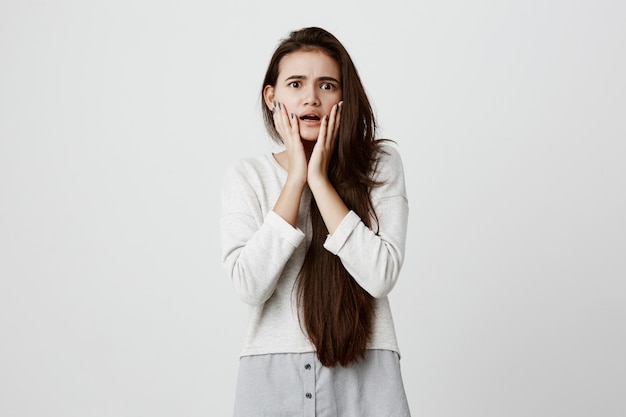 Emotional worried brunette woman holding hands on her cheeks, opening mouth, feeling perplexed and frustrated after she left home without having unplugged iron. Human emotions and feelings