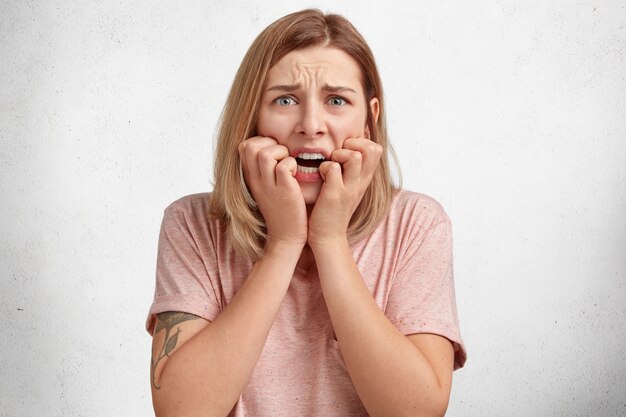 Emotional worried frightened young beautiful woman has puzzled expression, feels very nervous before passing exam, bites nails and looks desperately at camera, isolated over white concrete wall.