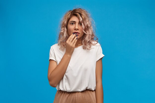 Emotional worried young woman accountant with stylish hairdo posing against blank blue  wall space, touching lips, being nervous