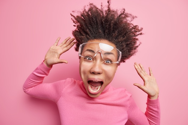 Free photo emotional young afro american woman screams loudly has funny face hair floating in air keeps hands raised wears transparent glasses turtleneck