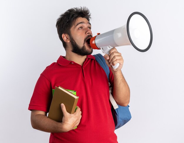 Emotional young bearded student guy in red polo shirt with backpack holding notebooks shouting to megaphone standing over white wall