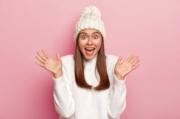 Emotional young Caucasian woman raises palms, keeps mouth opened, has fun, dressed in winter white clothes, poses over pink background.