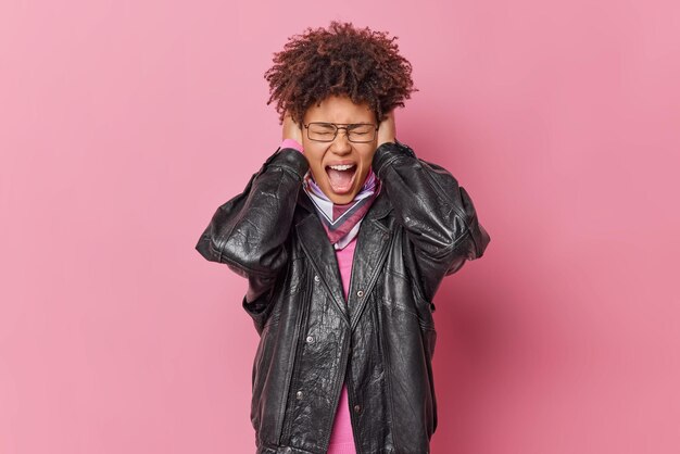 Emotional young curly haired woman avoids too loud noise exclaims loudly covers ears with hands wears spectacles and leather jacket isolated over pink studio background. Oh no how noisy is there