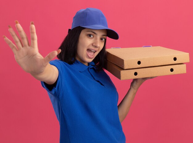 Эмоциональная молодая доставщица в синей униформе и кепке держит коробки для пиццы, делая жест с открытой ладонью, стоя над розовой стеной