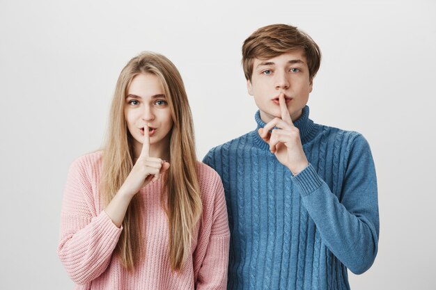 Emotional young European man and woman with blonde hair dressed in pink and blue sweaters holding fingers at lips