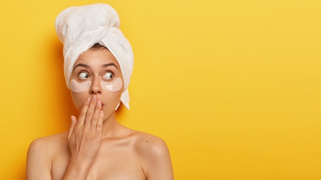 emotional young lady covers mouth with hand, looks with bugged eyes, has bated breath, being topless, looks away, cares about delicate skin under eyes