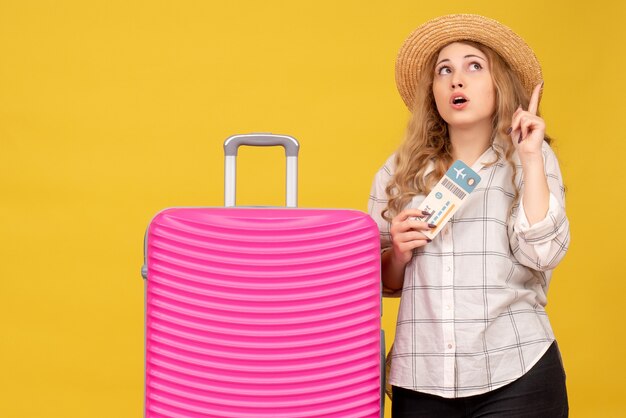 Emotional young lady wearing hat showing ticket and standing near her pink bag pointing up