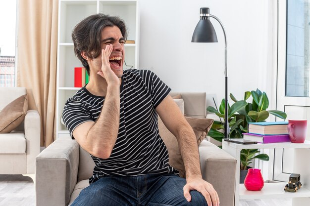 Emotional young man in casual clothes shouting loudly or calling someone with hand near mouth sitting on the chair in light living room