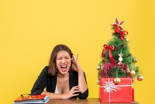 Emotional young woman closing her eyes sitting at a table near decorated Christmas tree at office on yellow 