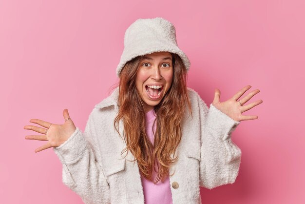 Emotional young woman raises palms and exclaims loudly being full of fun foolishes around wears stylish panama and fur jacket isolated over pink background. Human reactions and happiness concept