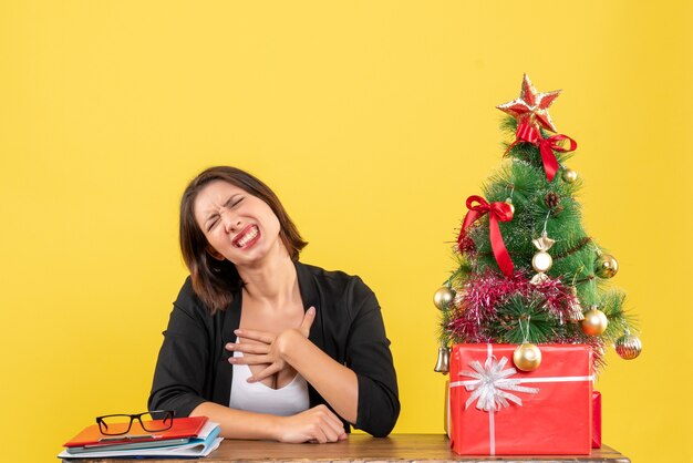 Emotional young woman sitting at a table near decorated Christmas tree at office on yellow 