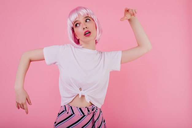 感情的な若い女性は、浮気する短いピンクのペルークを着ています。明るい壁に面白いダンスをかつらでうれしい白人の女の子